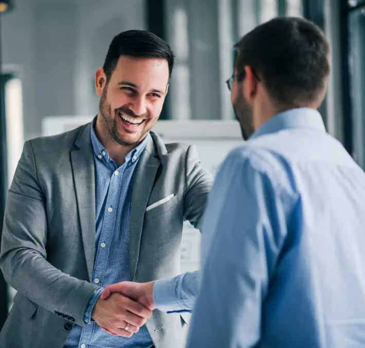 Two men in business attire are shaking hands indoors, with one smiling. The setting appears to be a professional or office environment.